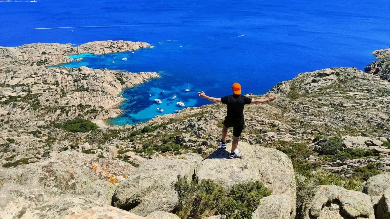 Uomo con braccia aperte ammira la splendida Cala Coticcio, con le sue acque turchesi e le barche all'ancora.