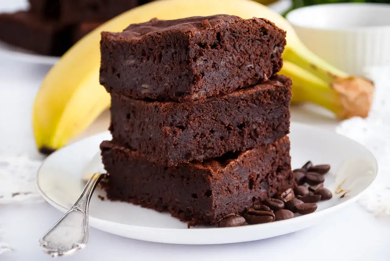 Stos pysznych brownie z bananami, ozdobiony ziarnami kawy, na talerzu z widocznym bananem w tle.