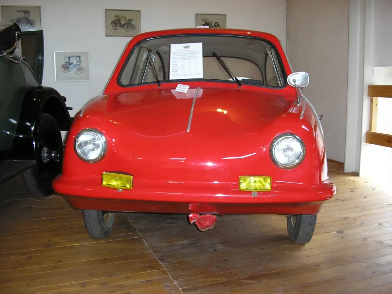 En r&ouml;d, liten bil, en Messerschmitt KR200, st&aring;r p&aring; ett tr&auml;golv. Fram king fulda.