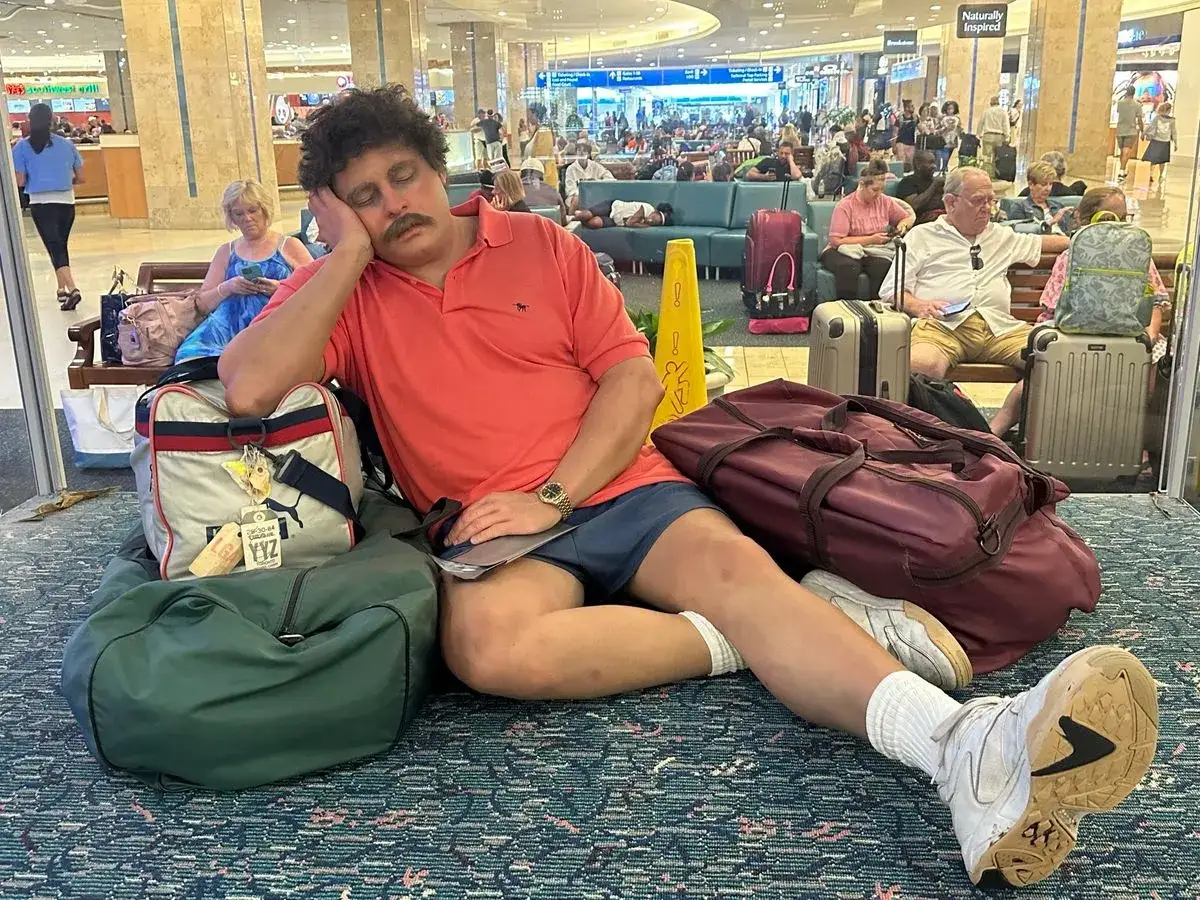 Zmęczony podróżnik z wąsami śpi na lotnisku, otoczony bagażami. Wygląda na to, że **jetlag** dał mu się we znaki.