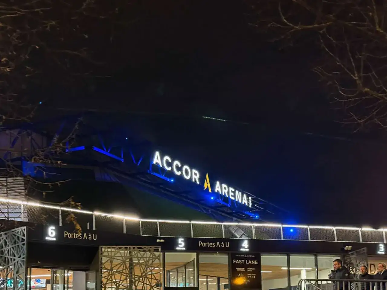 La nuit tombe sur l'Accor Arena, illuminée de néons bleus. Les portes d'entrée sont visibles, prêtes à accueillir les spectateurs.