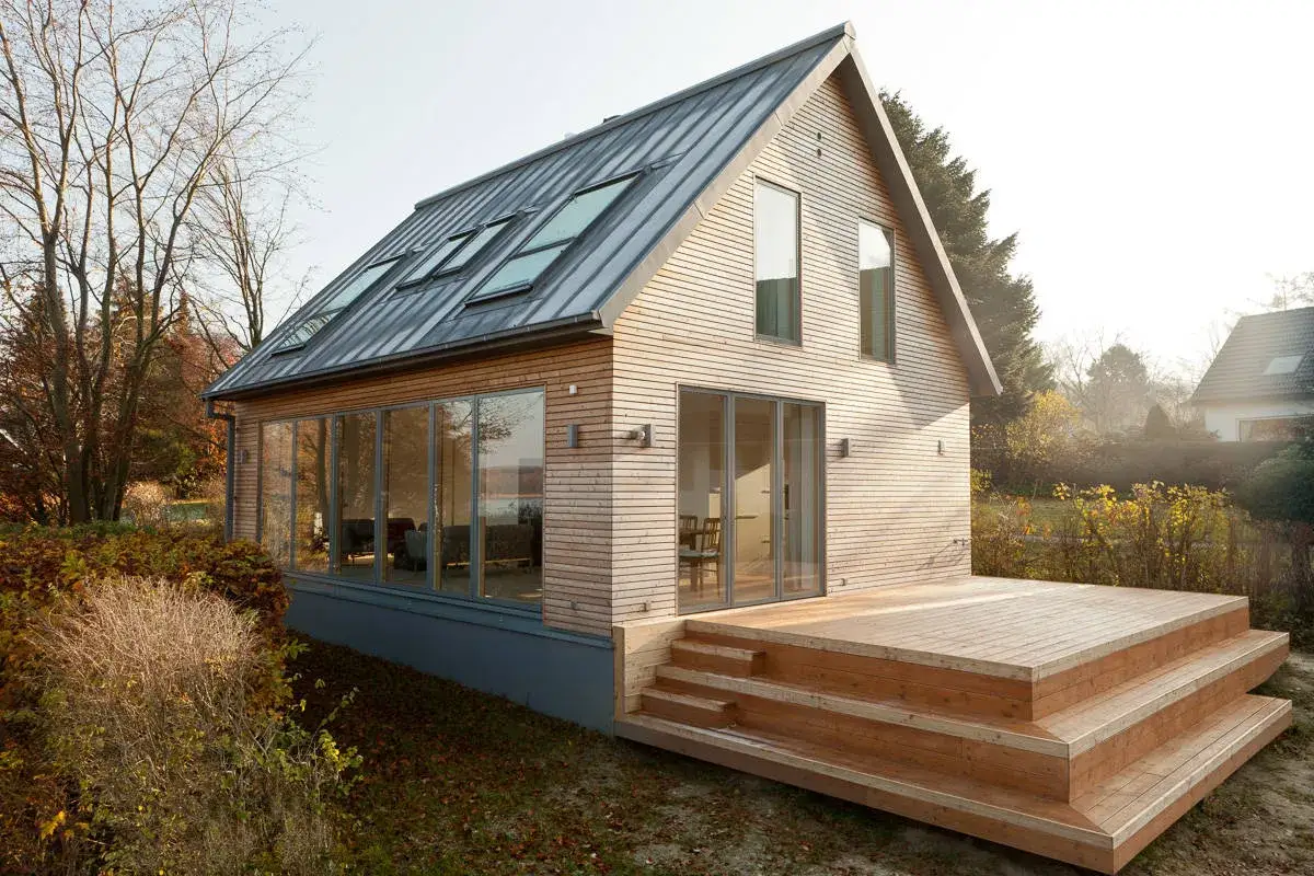Modernes Holzhaus mit gro&szlig;en Fenstern und Dachgauben. Eine Holzterrasse mit Stufen l&auml;dt zum Verweilen ein. Ein Beispiel, wie man ein low budget haus bauen kann.