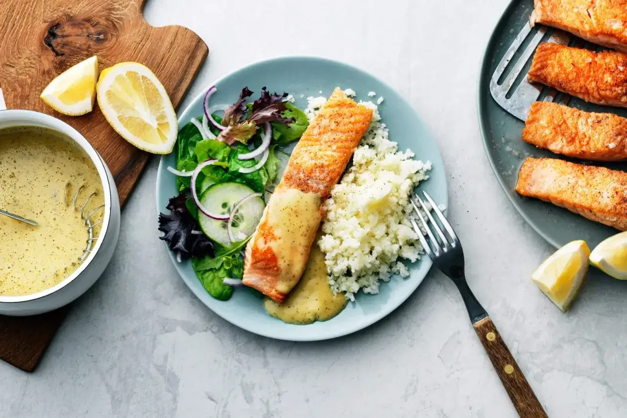 En tallrik med laxfil&eacute;, blomk&aring;lsmos och en fr&auml;sch sallad. En sk&aring;l med kr&auml;mig s&aring;s till laxen st&aring;r bredvid, med citronklyftor som dekoration.