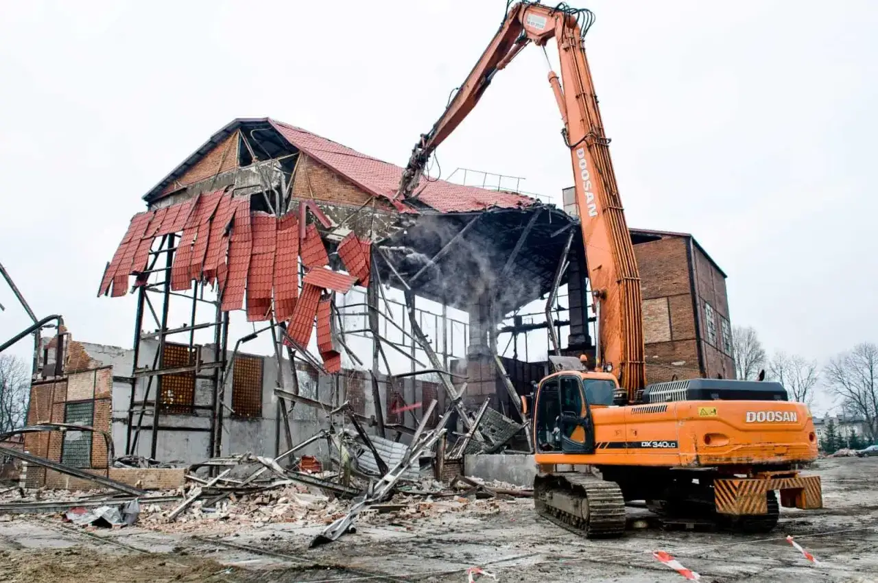 Pomarańczowa koparka do wyburzeń pracuje przy rozbiórce zniszczonego budynku, krusząc dach i ściany.