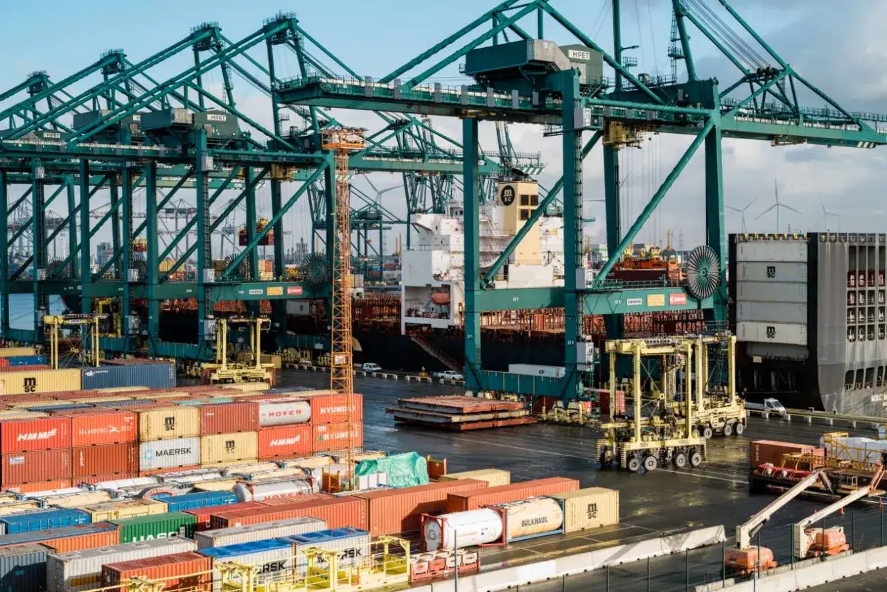 Massive green cranes dominate the Antwerp port, loading containers onto a colossal ship. Wind turbines spin in the distance.
