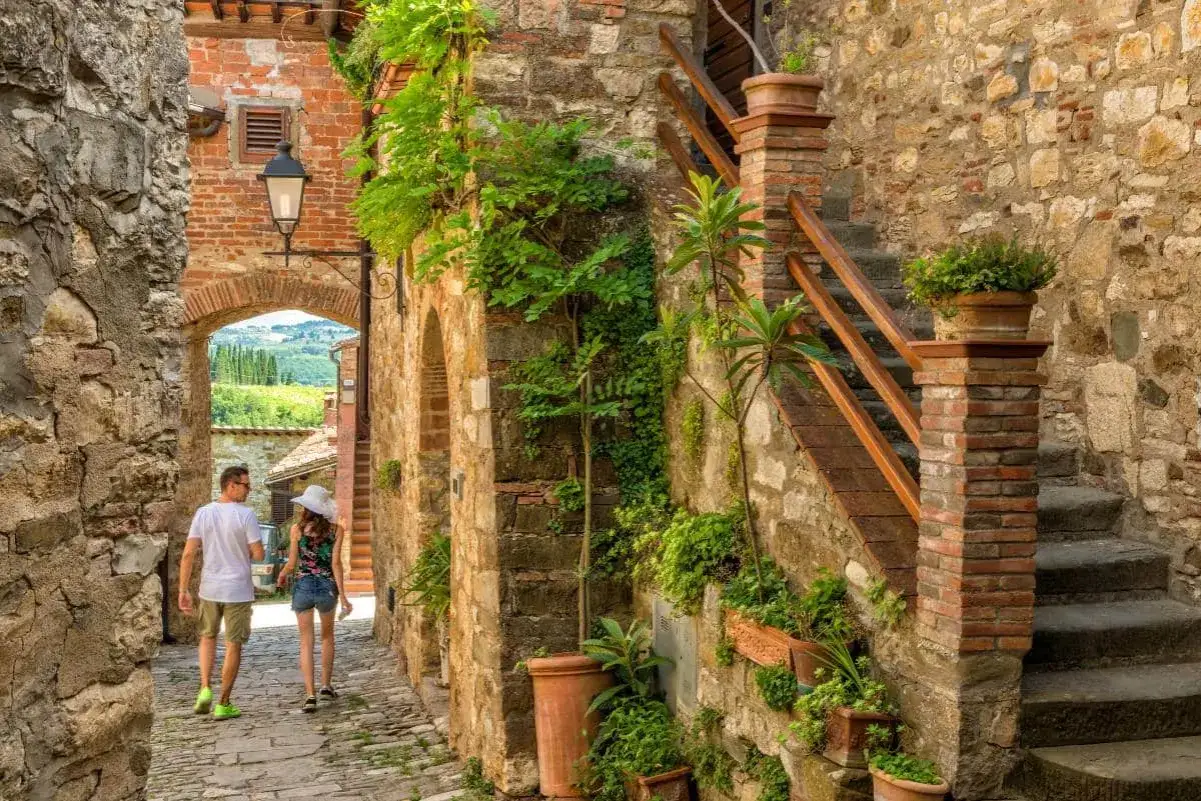 Coppia passeggia in un vicolo di pietra, tra i borghi da visitare, con piante rampicanti e un panorama verde all'orizzonte.