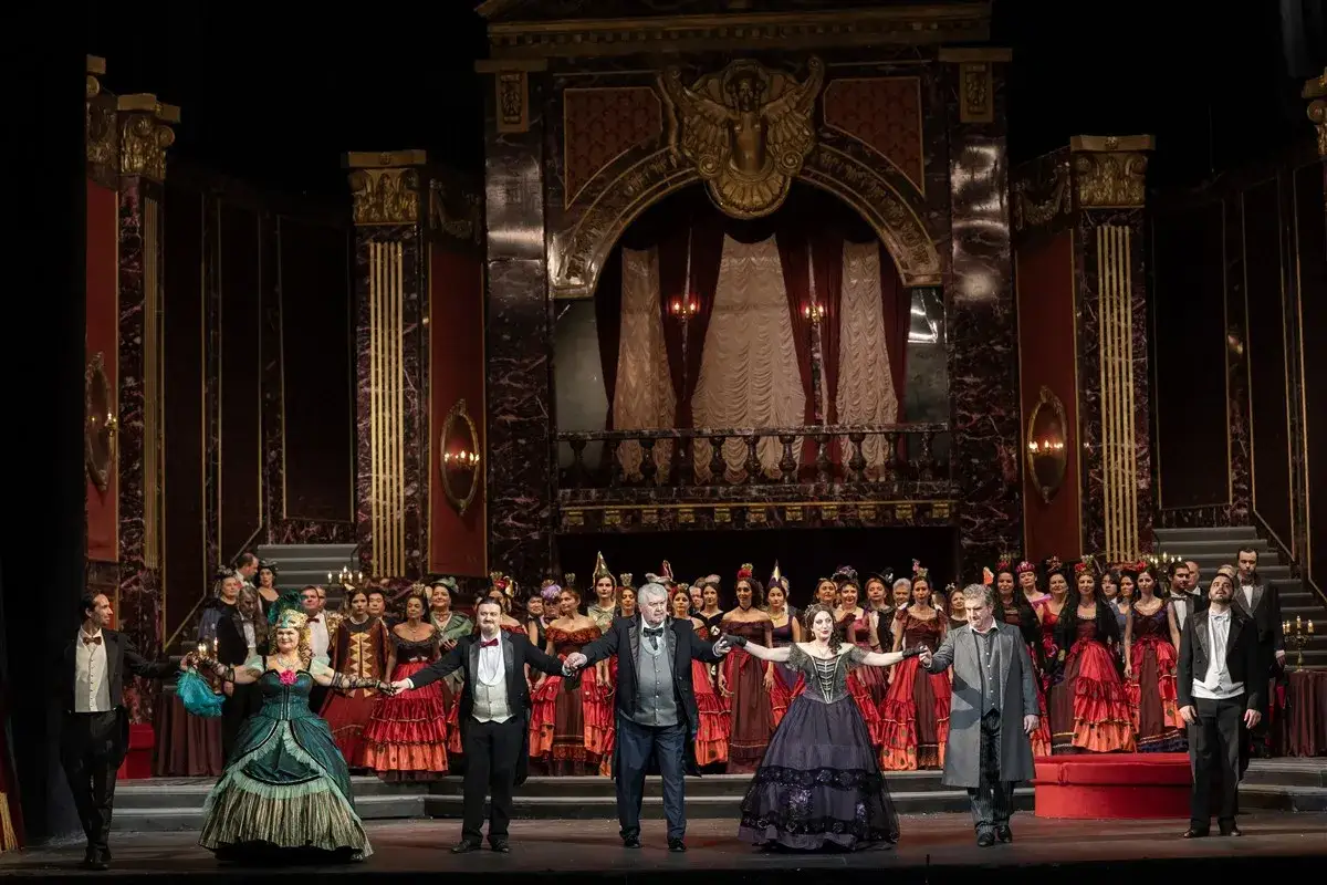 Aktorzy w strojach z epoki na scenie operowej. W tle bogata scenografia, przypominająca pałacowe wnętrza. Wiele tytułów oper Verdiego mogłoby się rozgrywać w tak wspaniałej scenerii.