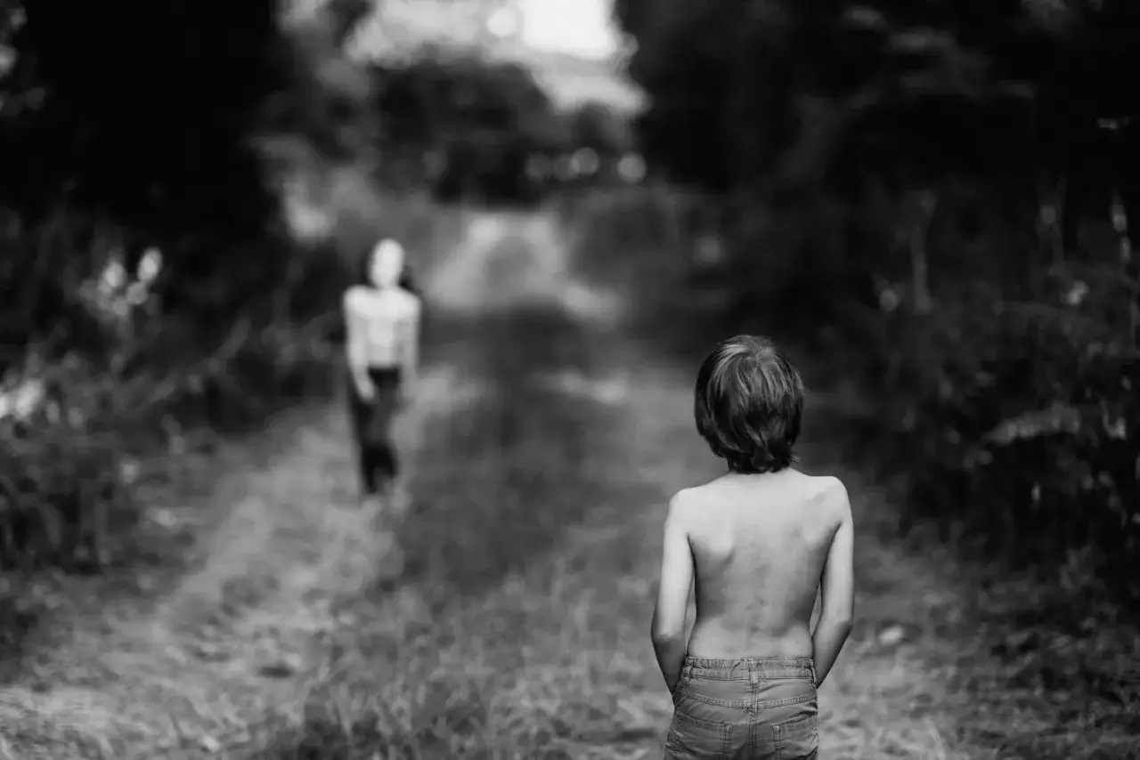 Bambino a torso nudo con le mani nelle tasche dei jeans, in un sentiero boscoso. Un'altra figura &egrave; sfocata in lontananza.