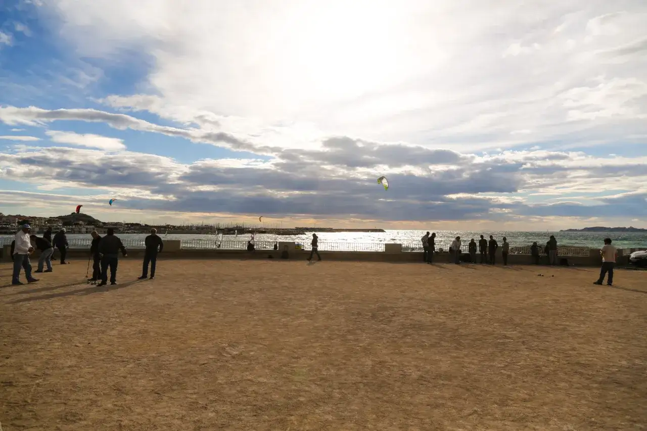 Des joueurs de p&eacute;tanque profitent d'une journ&eacute;e ensoleill&eacute;e au bord de la mer, avec des kitesurfeurs en arri&egrave;re-plan.