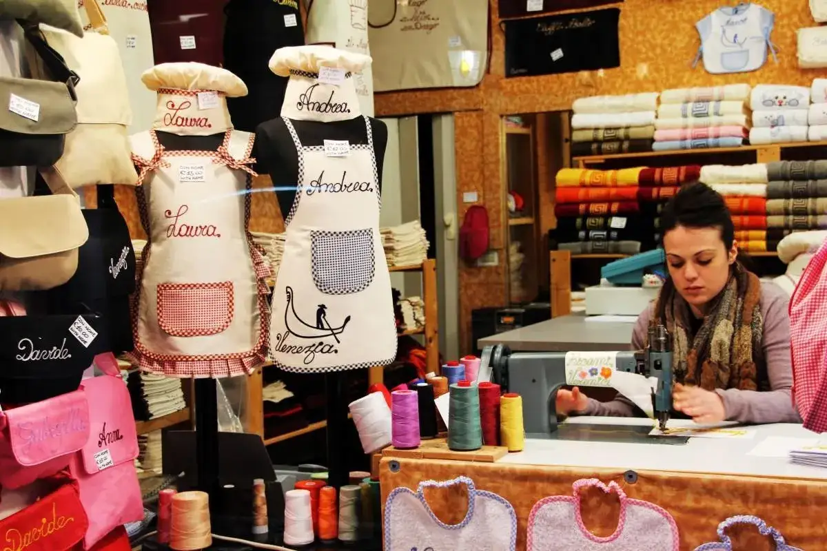 En esta mercería, delantales personalizados con nombres como Laura y Andrea, y bolsos bordados. Una mujer cose en una máquina de coser.