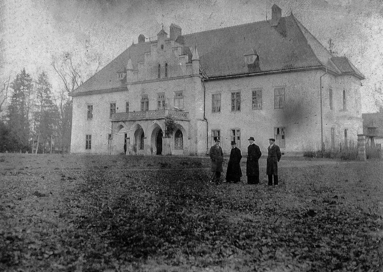 Stare zdjęcie przedstawia dwór w Łodygowicach. Przed budynkiem stoi grupa osób w strojach z epoki.