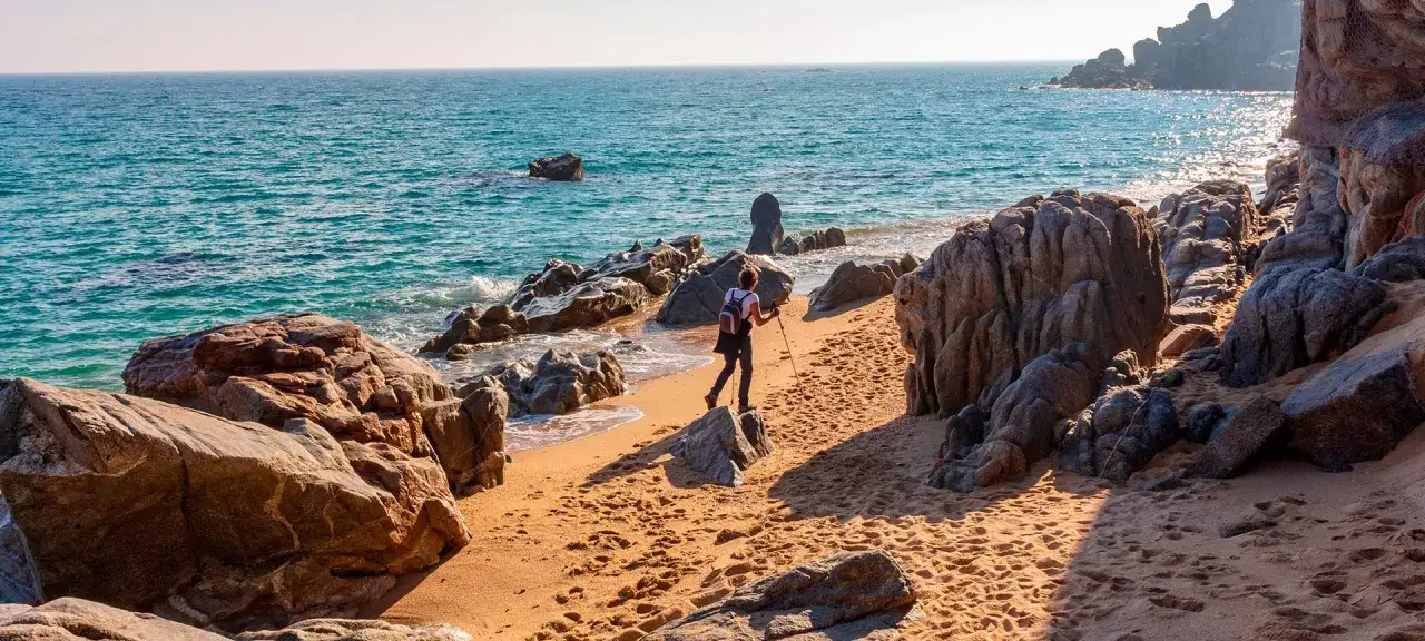 Kobieta z kijkami trekkingowymi idzie piaszczystą plażą wzdłuż skalistego wybrzeża. To piękny fragment szlaku cami de ronda.