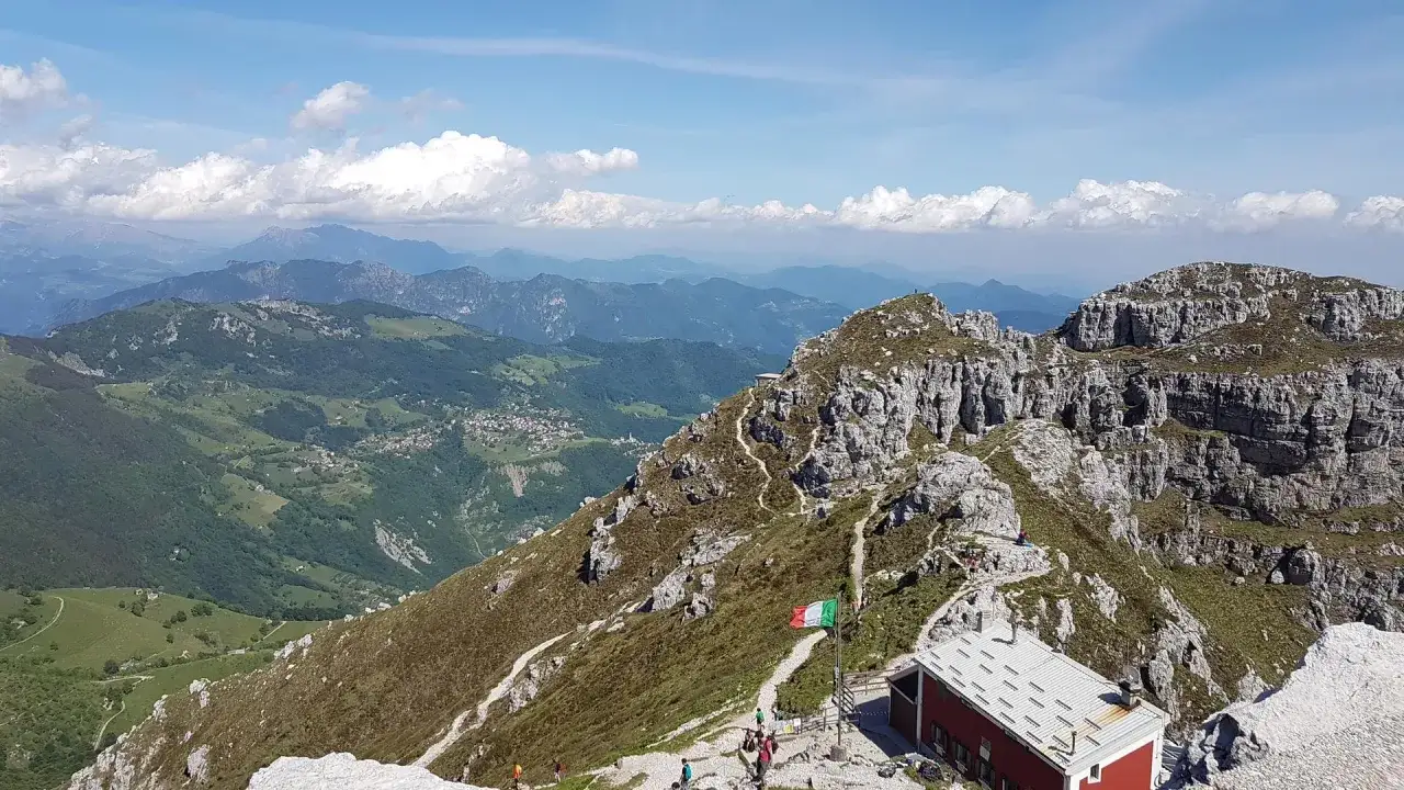 Il rifugio Lecco, con la bandiera italiana sventolante, domina un paesaggio montano mozzafiato. Sentieri serpeggiano tra rocce e prati verdi, con un villaggio in lontananza.