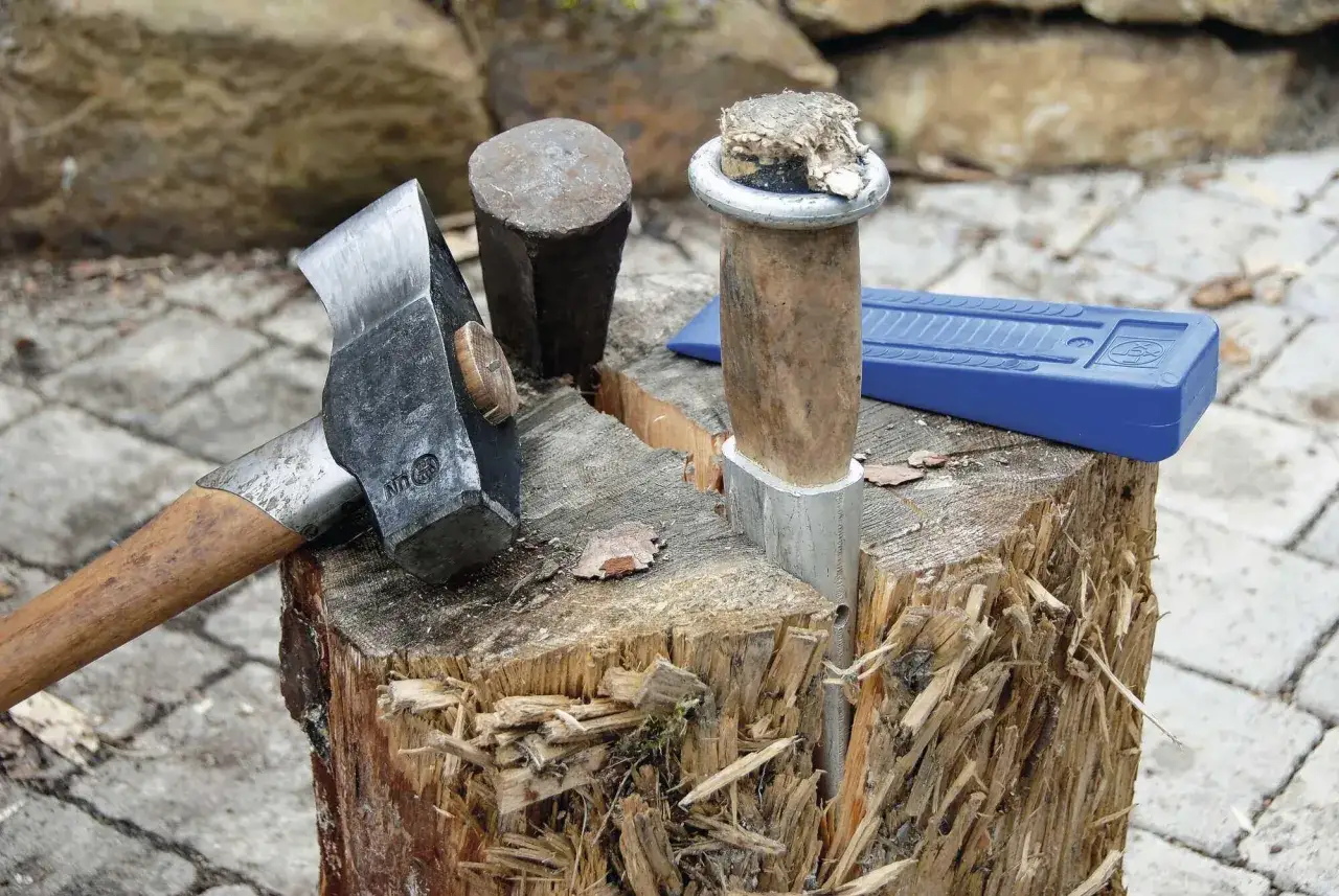Werkzeuge zum Holz spalten: Beil, Spalthammer, Keil und ein gespaltenes Holzstück auf einem Baumstumpf.
