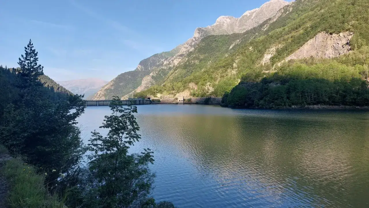 Embalse sereno en Alta Ribagorza, rodeado de monta&ntilde;as verdes y picos rocosos bajo un cielo azul.
