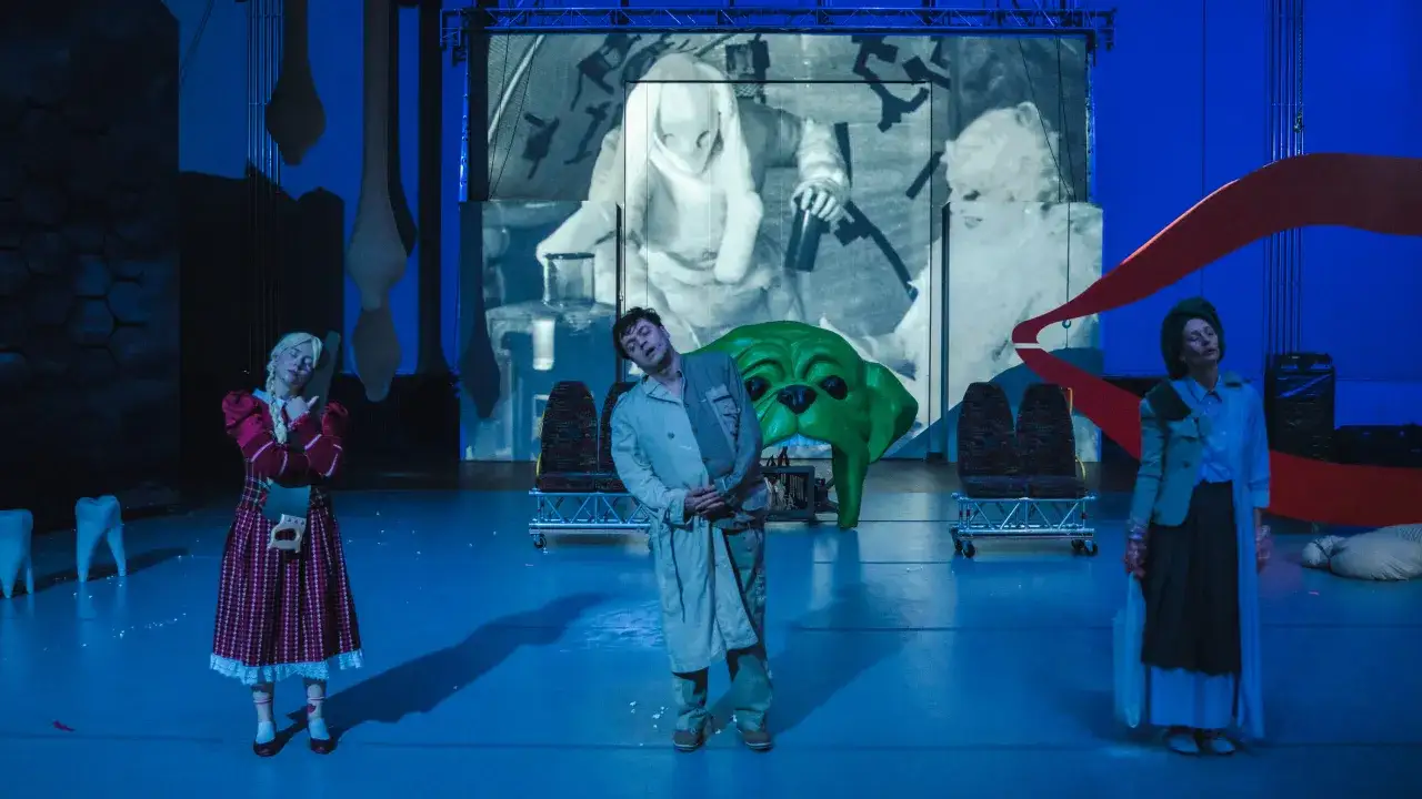 Scena teatralna z elementami surrealizmu, przypominająca świat z prozy kafki. Trzy postacie na niebieskiej scenie, w tle projekcja i groteskowe rekwizyty.