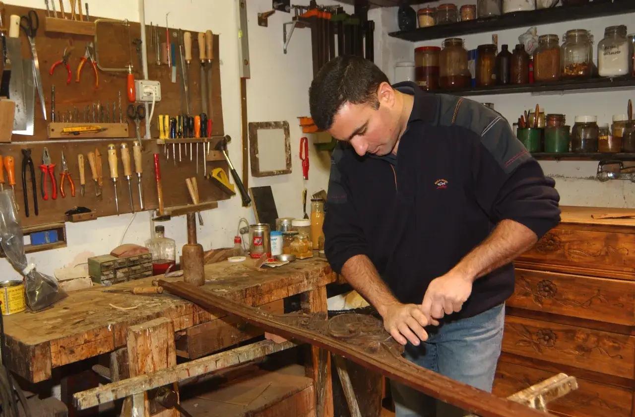 Un artigiano lavora il legno con precisione, circondato da attrezzi e materiali. La sua abilit&agrave; nel lavorare il legno &egrave; evidente nel dettaglio del pezzo che sta rifinendo.