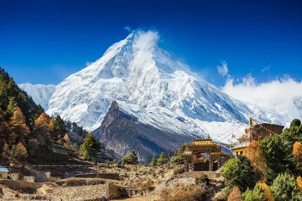 Majestatyczny ośnieżony szczyt góruje nad wioską z buddyjską bramą i kolorowymi flagami. Widok zapierający dech w piersiach podczas Manaslu Circuit Trek.
