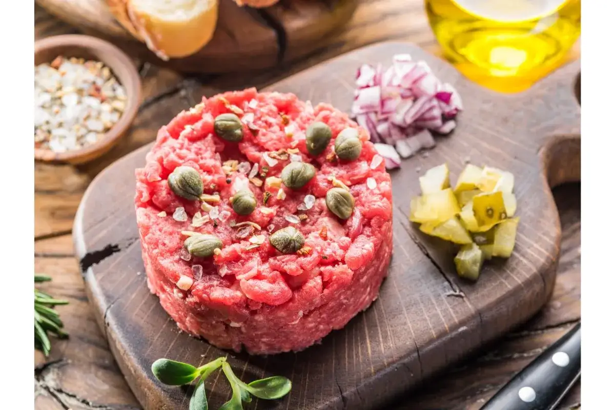 Un d&eacute;licieux tartare de b&oelig;uf gastronomique, garni de c&acirc;pres et de sel, accompagn&eacute; d'oignons rouges et de cornichons sur une planche en bois.