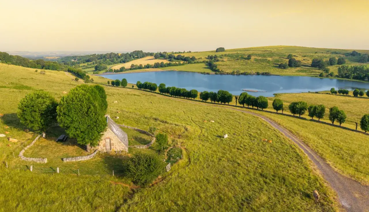 Vue aérienne d'un petit bâtiment en pierre près d'un lac, sur fond de collines verdoyantes. Un chemin serpente à travers les pâturages où paissent des vaches. Paysage typique du tour de l'Aubrac.