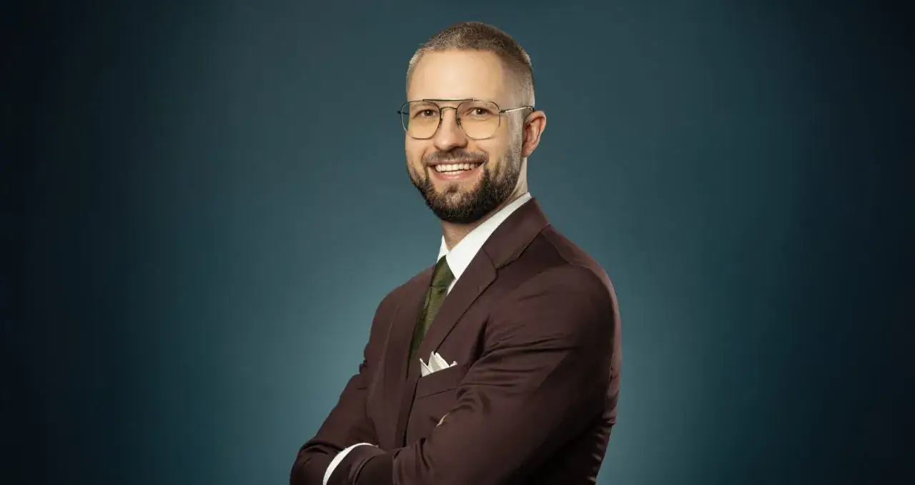 Uśmiechnięty mężczyzna w garniturze i okularach, ze skrzyżowanymi rękami, na ciemnym tle.