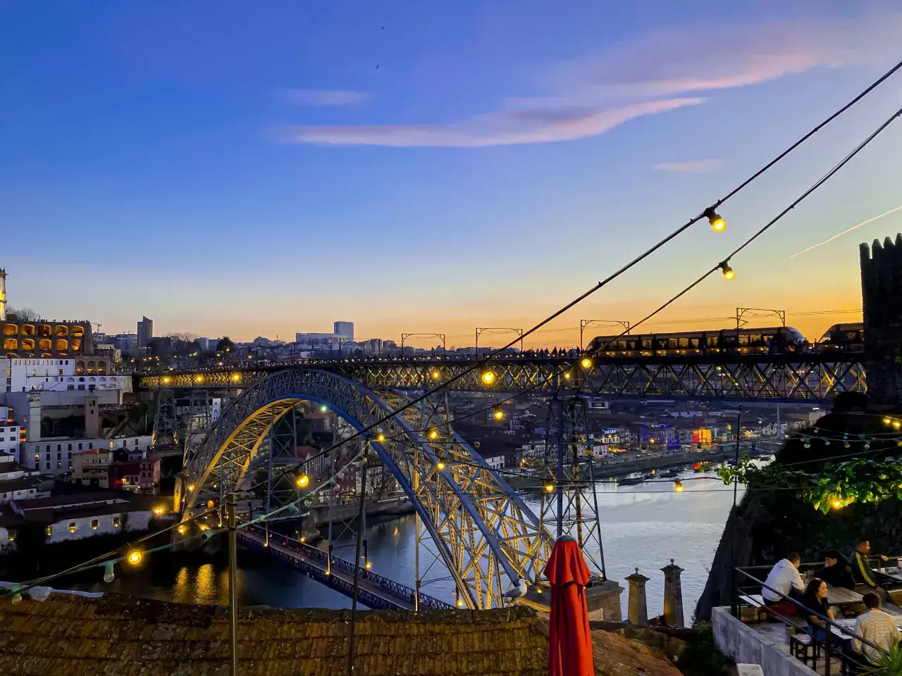 Ponte Dom Lu&iacute;s I illuminato al tramonto, con un treno in transito e luci che creano un'atmosfera da porto vita notturna.