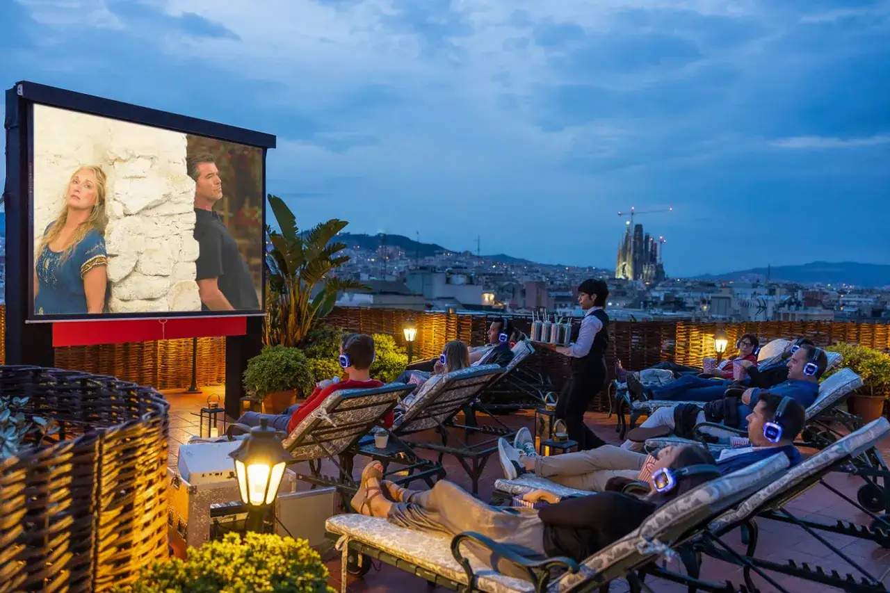 Cinema all'aperto su un tetto di **night Barcelona**. Persone rilassate su lettini, con cuffie, guardano un film con la Sagrada Familia sullo sfondo.