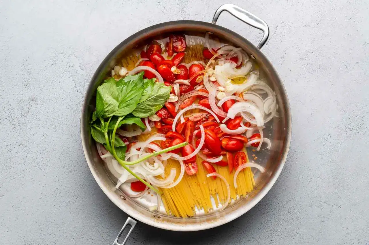 Zutaten für eine One-Pot-Pasta: Spaghetti, Kirschtomaten, Zwiebelringe und frisches Basilikum in einer Pfanne.