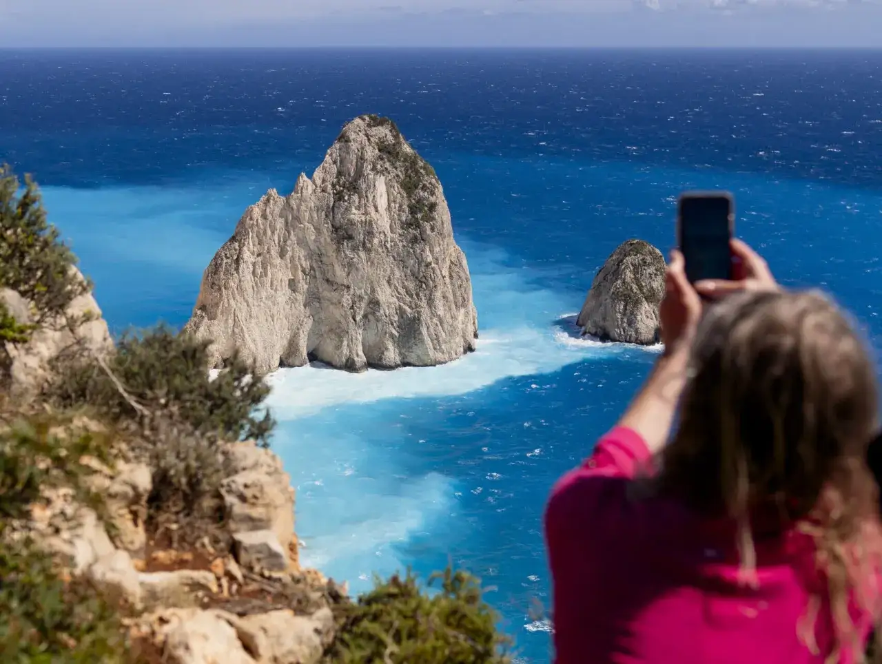 Widok na turkusowe morze i skaliste wysepki koło miasta Zakynthos. Kobieta robi zdjęcie telefonem.