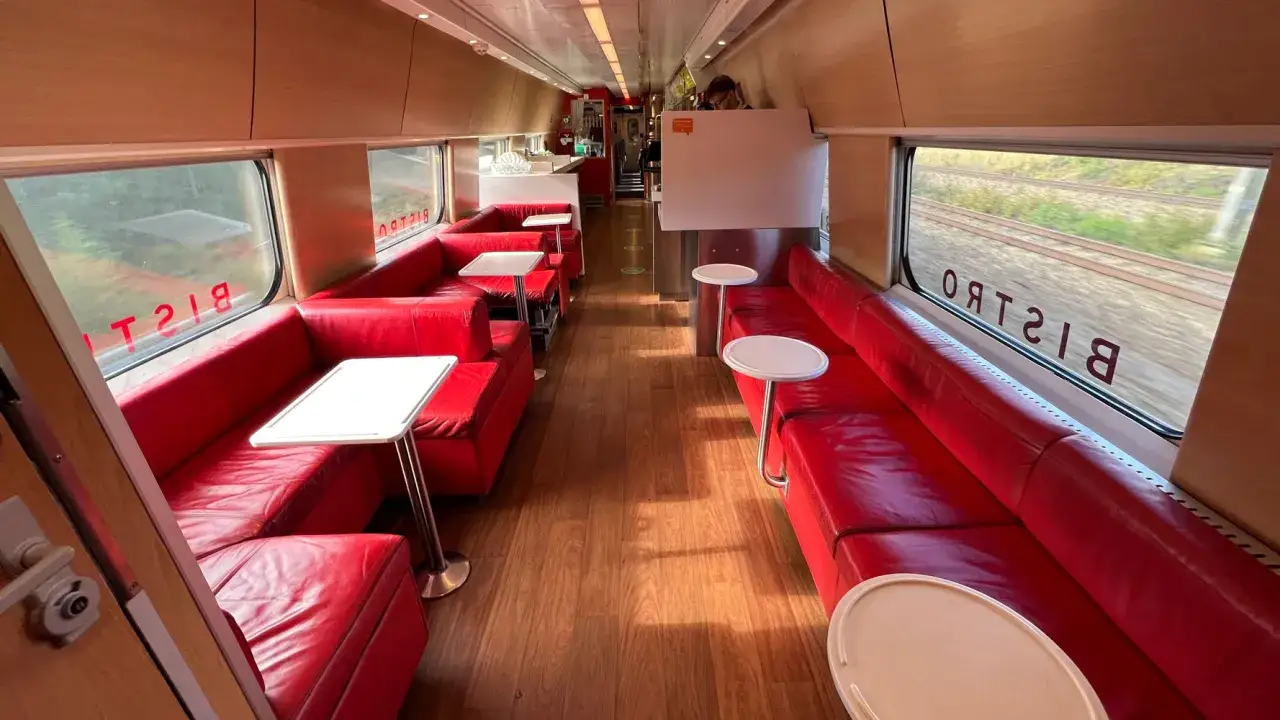 Innenraum eines SJ Intercity Bistrowagens mit roten Ledersitzen und Holzboden. Drau&szlig;en vorbeiziehende Landschaft.
