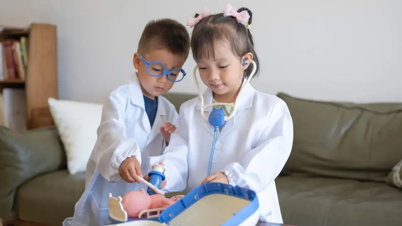 Dos ni&ntilde;os de 4 a 5 a&ntilde;os, con batas blancas, juegan a ser doctores. Uno examina un mu&ntilde;eco mientras el otro escucha con un estetoscopio.