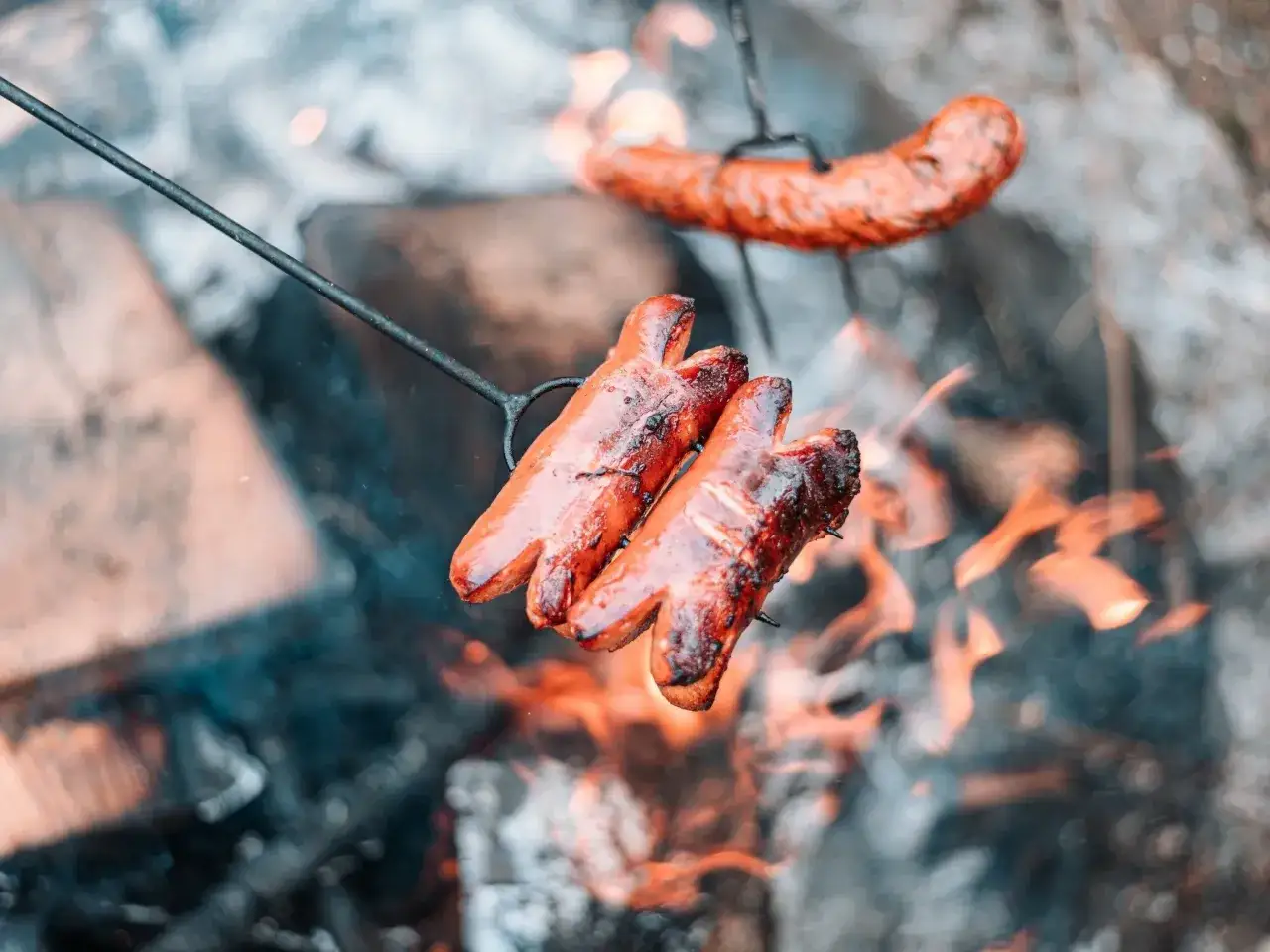 Trzy soczyste kiełbaski na ognisko pieką się nad płomieniami. Jedna kiełbasa w tle również apetycznie się rumieni.