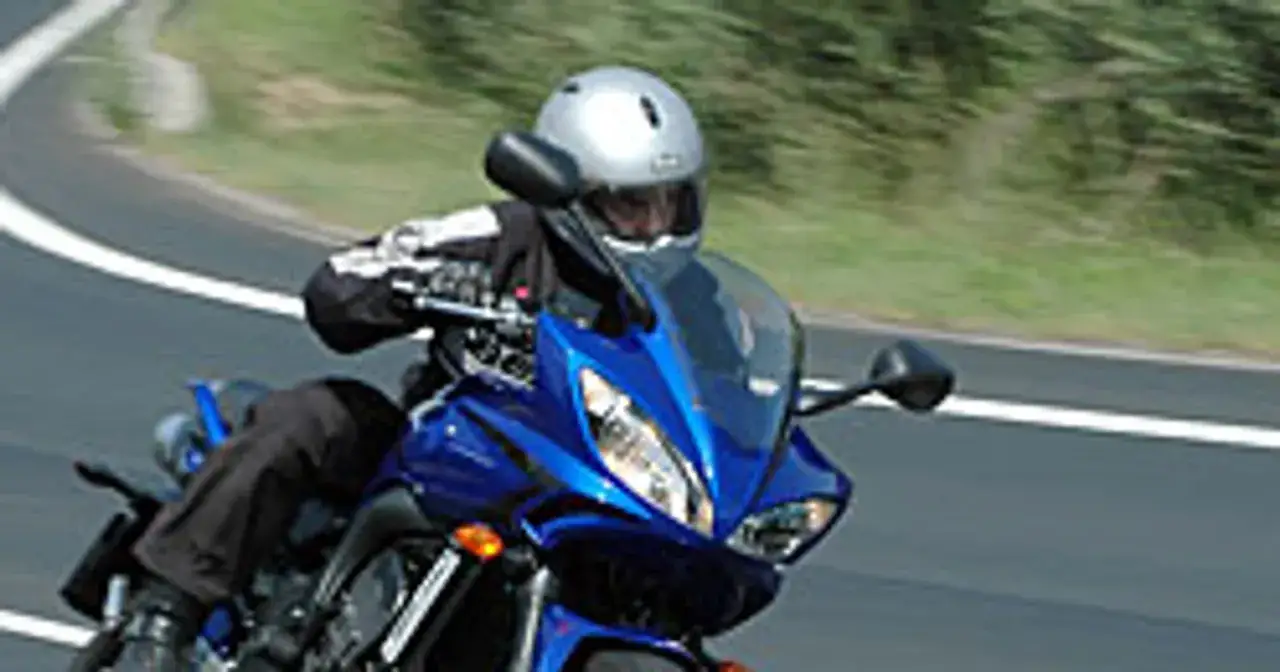 Motocyklista w srebrnym kasku i czarnej kurtce pochyla się nad niebieskim fazerem, pokonując zakręt na torze.