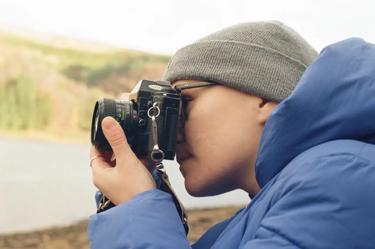 A person wearing a grey beanie and blue jacket takes a photo with a Nikon FE camera, capturing the landscape.