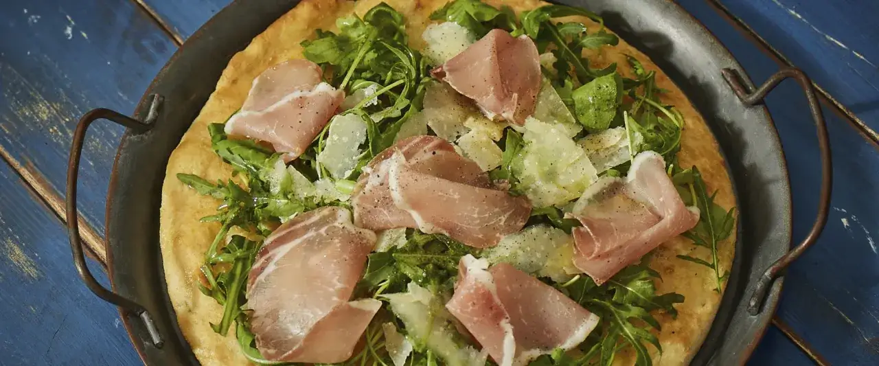En pizza med bresaola, ruccola och parmesan på en rustik plåt.