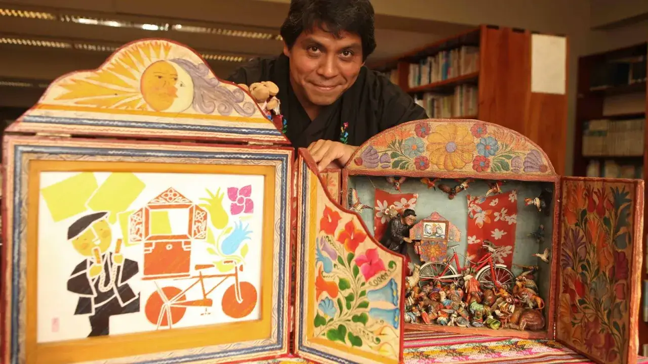 Hombre sonriente muestra un kamishibai casero, un teatro de t&iacute;teres con escenas detalladas y coloridas, lleno de figuras y motivos folcl&oacute;ricos.