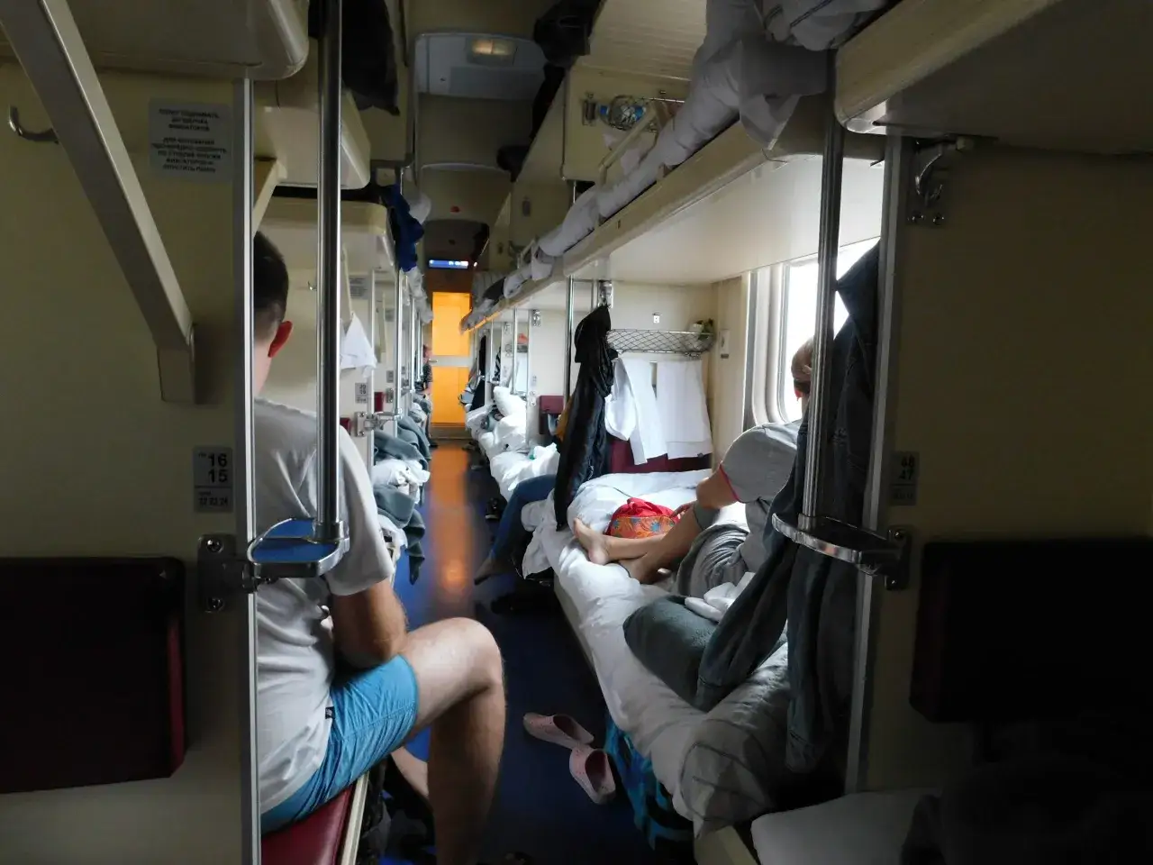 Wnętrze wagonu kolejowego, podróżni odpoczywają na kuszetkach. Długi korytarz, typowy dla kolej transsyberyjska.