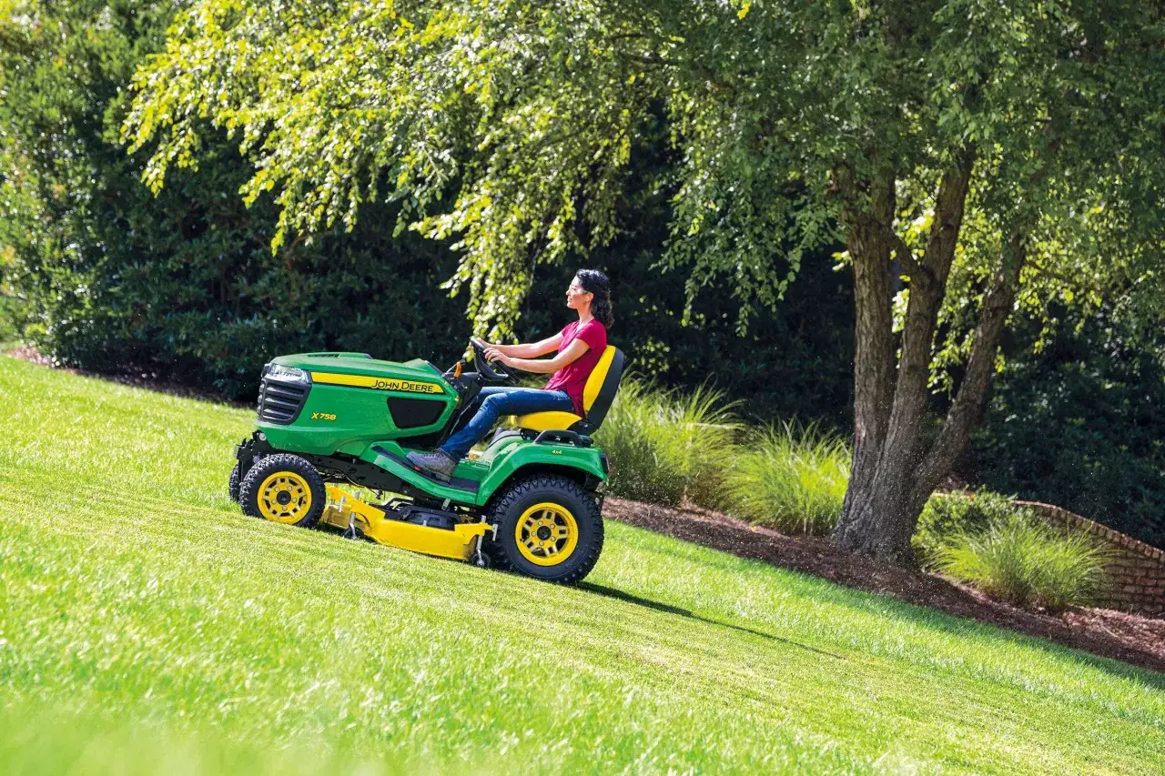 Kobieta w okularach prowadzi zieloną kosiarkę do jeżdżenia John Deere po pochyłej, zielonej trawie, podziwiając piękny, słoneczny dzień.