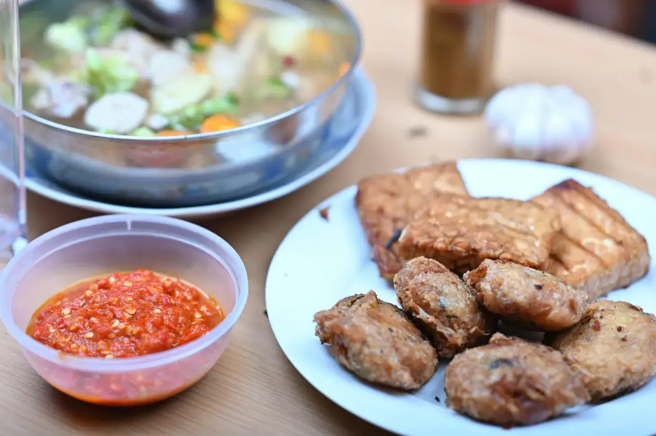 Pikantny sos sambal w miseczce obok talerza z smażonym tofu i kotlecikami. W tle zupa.