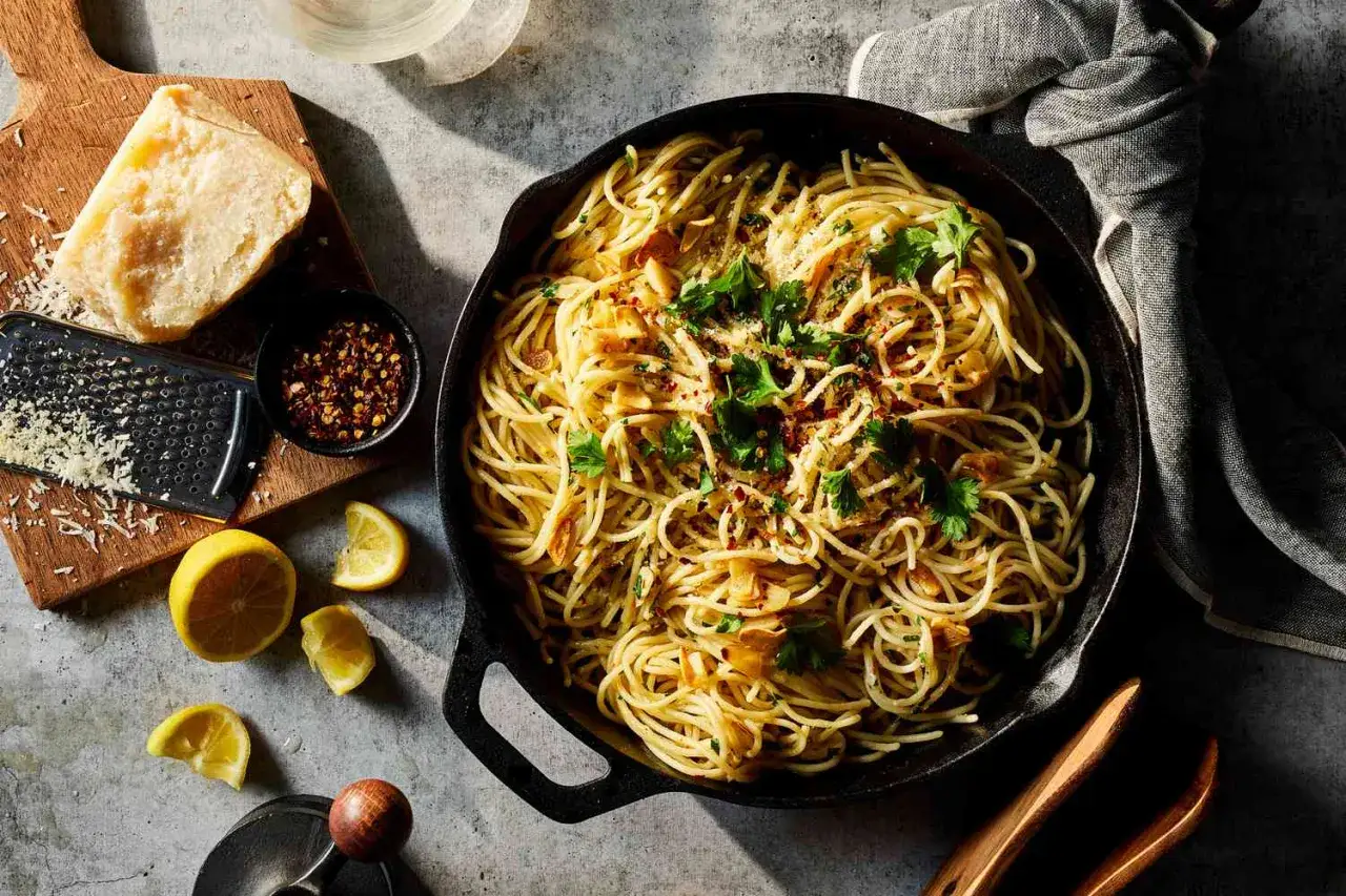 Un plato humeante de spaghetti aglio e olio, adornado con perejil fresco y hojuelas de chile rojo. A su lado, limones cortados, un trozo de queso parmesano y un rallador.