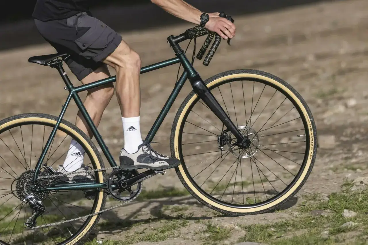 Mężczyzna na rowerze typu gravel pokonuje kamienistą ścieżkę. Zielona rama, szerokie opony i sportowe obuwie sugerują gotowość na każdą nawierzchnię.