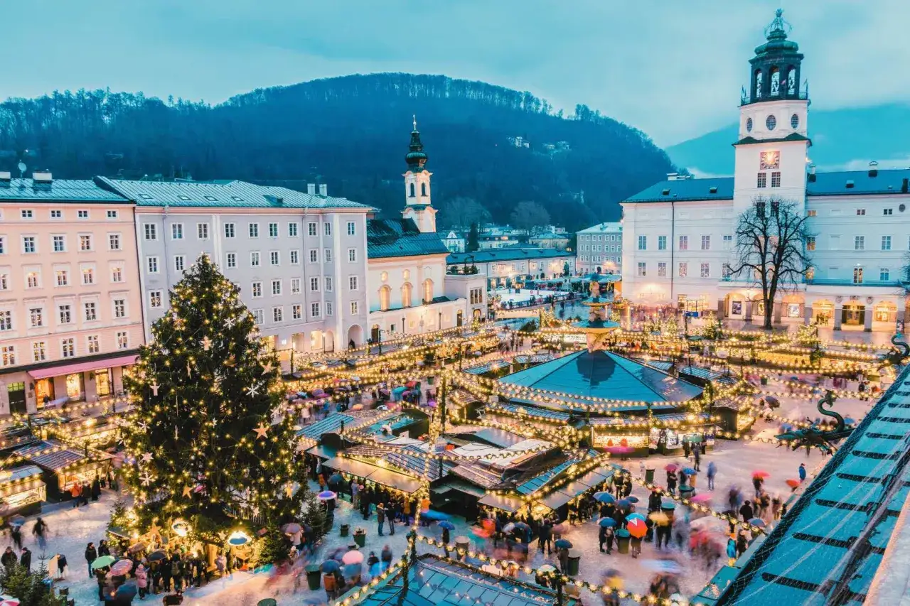A festive Salzburg Austria Christmas market glows with lights, a giant tree, and bustling crowds under umbrellas.