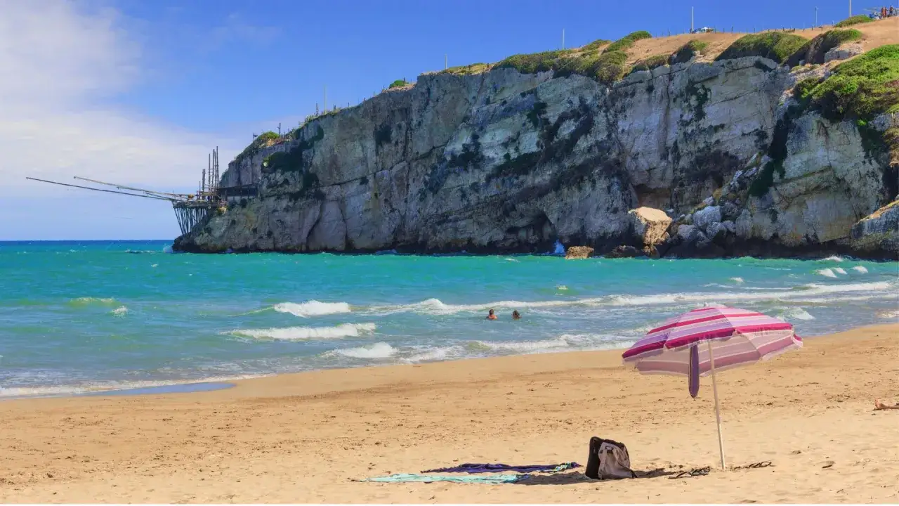 Spiaggia dorata con ombrellone a righe, mare turchese e scogliera con trabucco, un luogo incantevole come la cala della pipa.