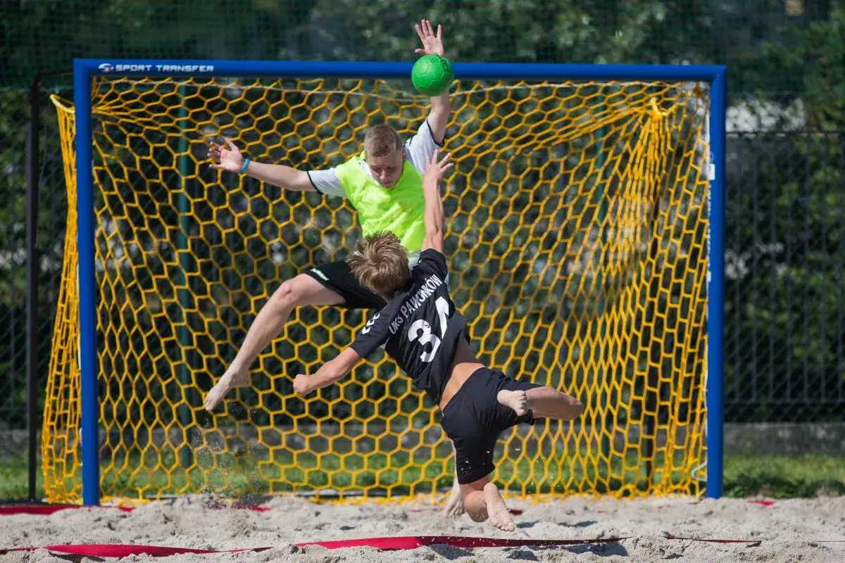 Dwóch zawodników w powietrzu walczy o piłkę podczas meczu beach handball. Jeden próbuje obronić bramkę, drugi atakuje.