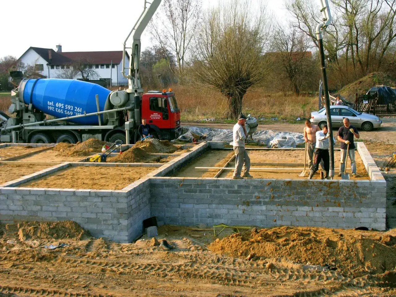 Budowa fundament&oacute;w: betoniarka i ekipa przygotowują teren. Czym obsypać fundamenty z zewnątrz? Piaskiem i ziemią.