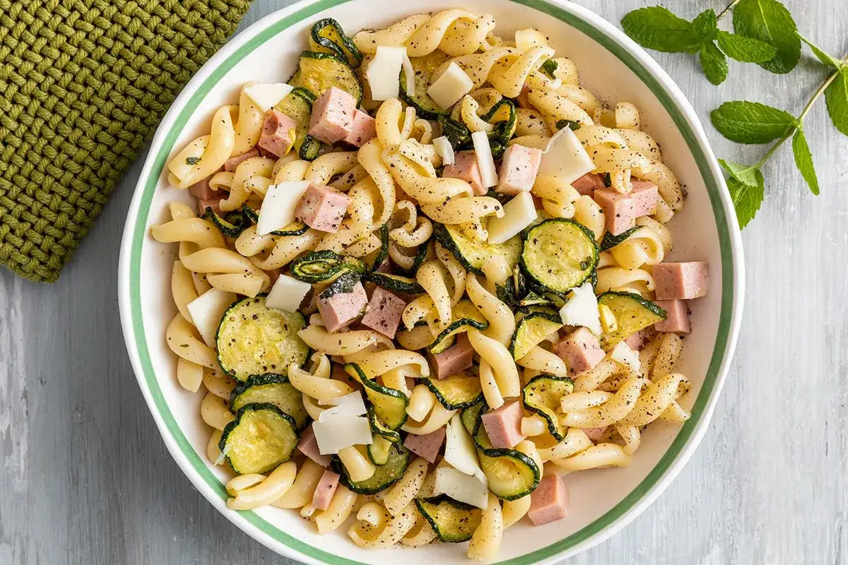 Un'insalata di pasta vegetariana con fusilli, zucchine grigliate, cubetti di formaggio e dadini di prosciutto cotto, condita con pepe nero.