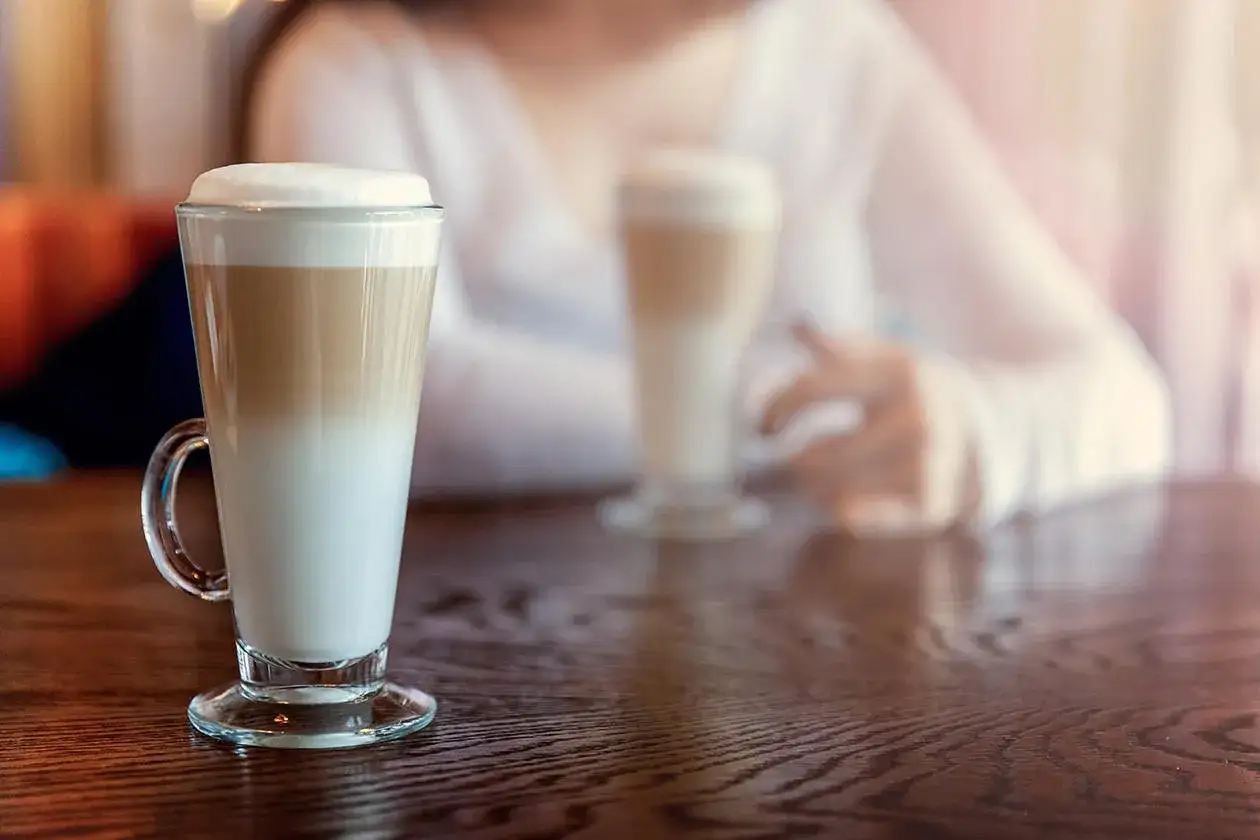 Ein Latte Macchiato mit Mandelmilch, perfekt geschichtet, steht auf einem Holztisch. Im Hintergrund eine Person.