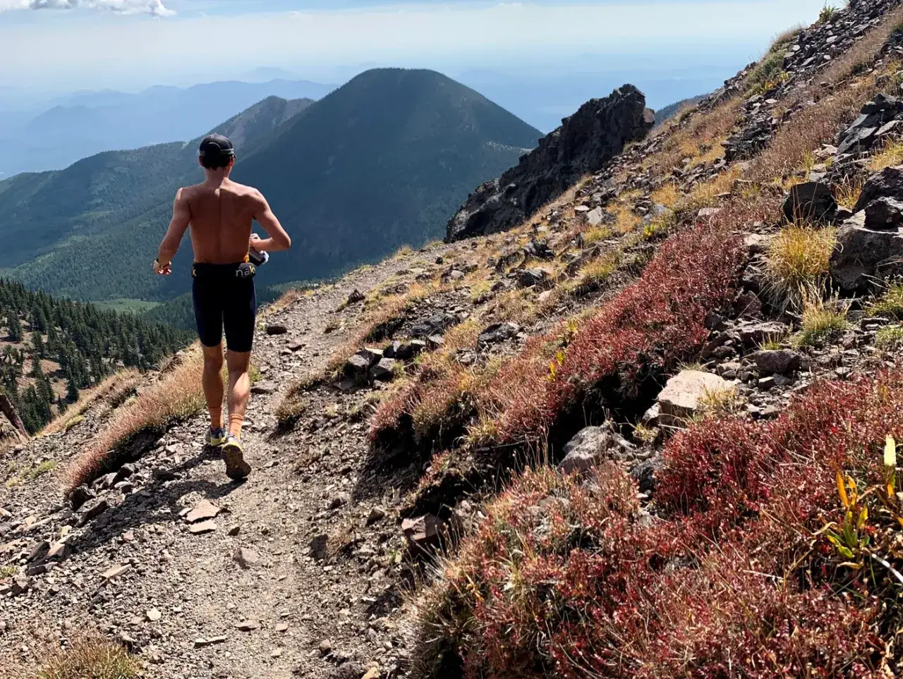 Un coureur sans torse d&eacute;vale un sentier **fractionn&eacute; trail** escarp&eacute;, entour&eacute; de v&eacute;g&eacute;tation rouge et de montagnes bleues &agrave; l'horizon.