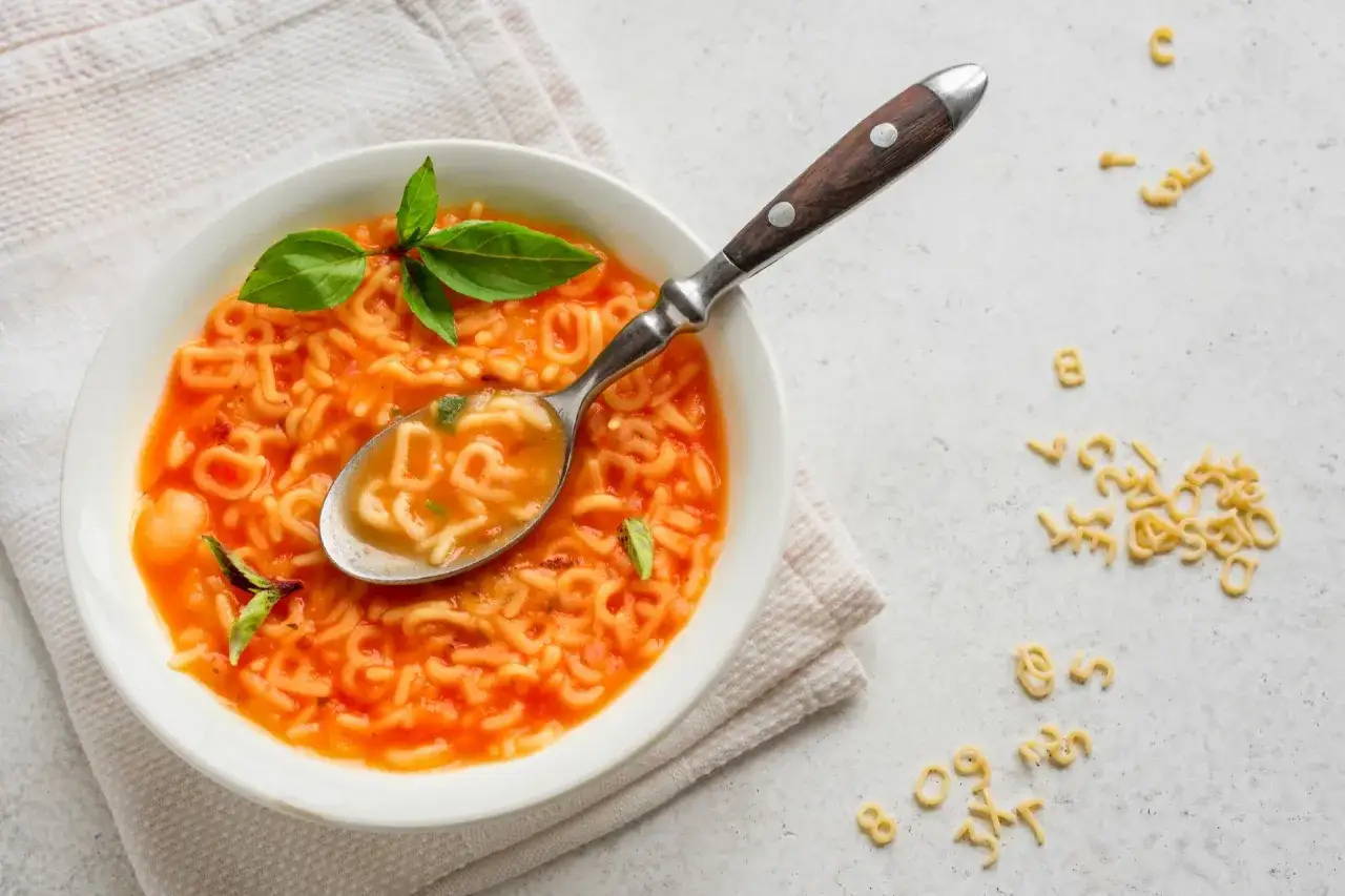 Zupa pomidorowa z pomidorów z puszki z makaronem w kształcie liter. Na łyżce widać literki, a obok rozsypane są kolejne.
