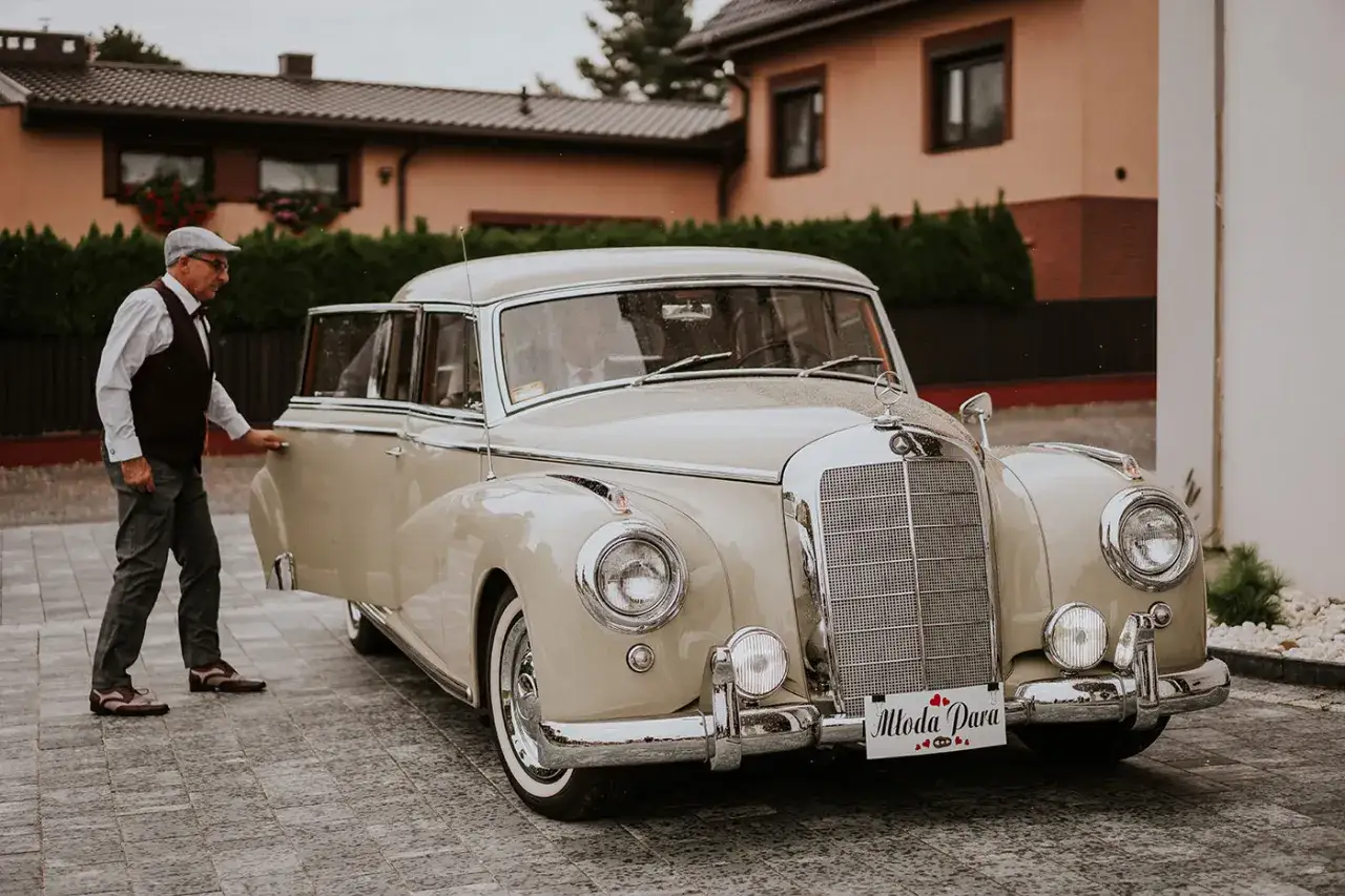Elegancki pan w kapeluszu otwiera drzwi zabytkowego Mercedesa. Zastanawiasz się, ile kosztuje wynajem samochodu do ślubu?